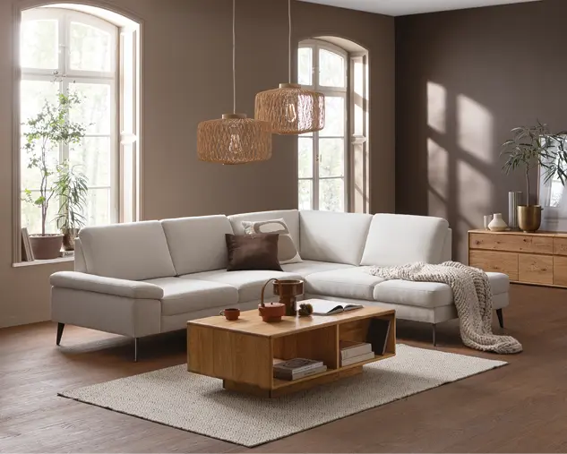 Beiges Ecksofa vor hohen Fenstern, davor niedriger Couchtisch aus Holz auf hellem Teppich, umgeben von Pflanzen und Sideboard im Wohnzimmerstil.